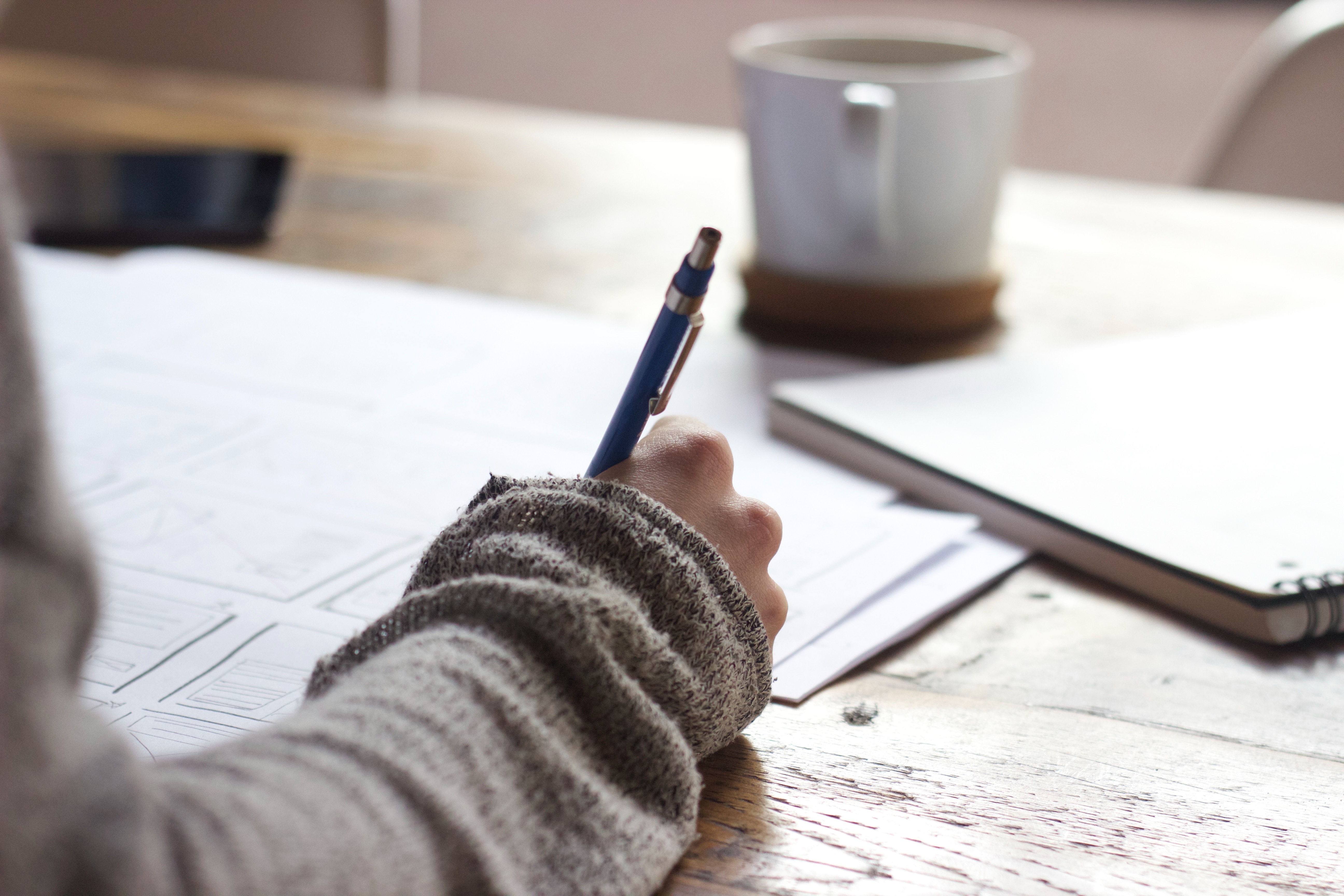 Close up of a hand writing with papers scattered on a table and cup of coffee in the background